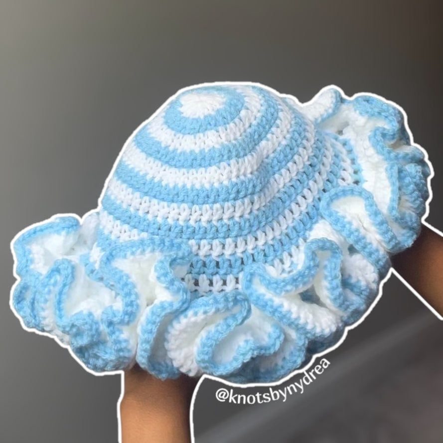 A handmade crochet sun hat with ruffled brim in light blue and white, held up by hands against a plain gray background.