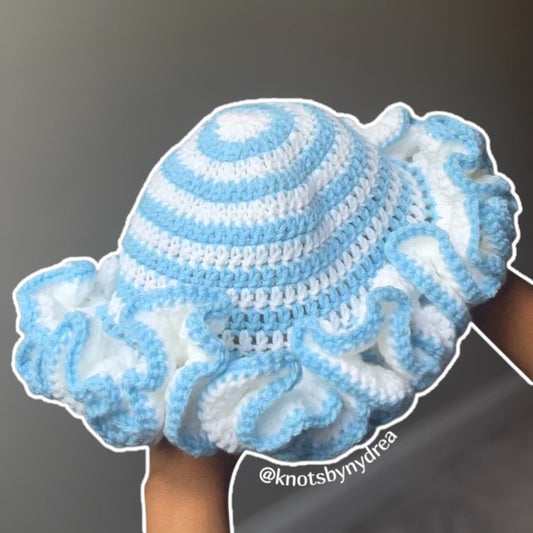 A handmade crochet sun hat with ruffled brim in light blue and white, held up by hands against a plain gray background.